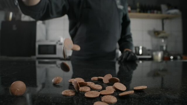 Chef chocolatier pours milk chocolate drops to the marble table in slow motion, raw materials for making sweets and chocolate, cooking the sweet desserts, making chocolate bars, Full HD 240fps Prores