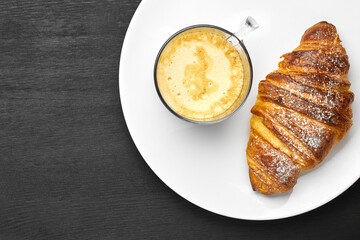 Top view of cappuccino and croissant on a plate on a black background with copy space. Concept of a tasty breakfast. French or italian pastry. 