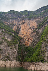 Guandukou, Hubei, China - May 7, 2010: Wu Gorge in Yangtze River: Huge beige brown rocky cliff set in wedge of mountain slopes with green foliage above green water under cloudscape.