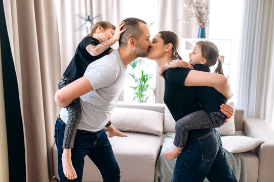 Parents and their two cute daughters. Young parents have fun at home with their beautiful funny daughters. Girls are on a backs of parents and dad and mom kiss each other