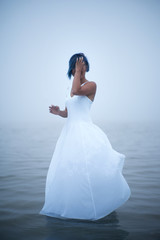 Fototapeta na wymiar Cute shy girl covers her face in white dress and blue hair looks as if she is drifting by water surrounded by mist and fog 
