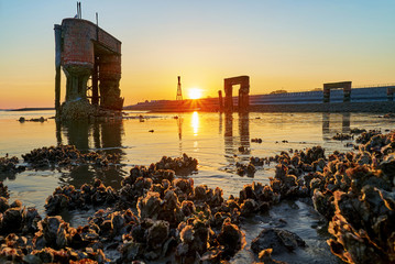 3 arches of the former landing bridge and silhouette of the historic lighthouse in Eckwarderhörne, district Wesermarsch, Germany during low tide against a beautiful sunset with oysters in foreground