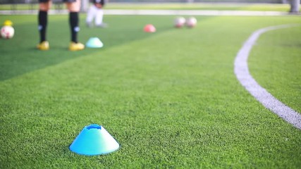 Selective focus to blue marker cone is soccer training equipment on green artificial turf with blurry kid players training background. Material for training class of football academy.