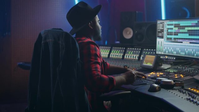 Energetic Male Audio Engineer / Producer Working in Music Recording Studio, Mixing Tracks on Control Desk and Software to Create Hit Song Track. Artist Musician Enter His Studio at Sits at Workdesk
