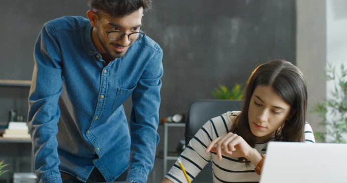 Young multiethnic male and female co-workers talking cheerfully and discussing work in office. Caucasian girl sitting at desk with laptop computer and Arab guy standing with tablet device as helping.