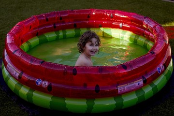 Caucasian girl plays with water in the plastic children's pool with watermelon colors. Child, boy enjoying water games in the summer heat. Exterior photo of a baby in a swimming pool.