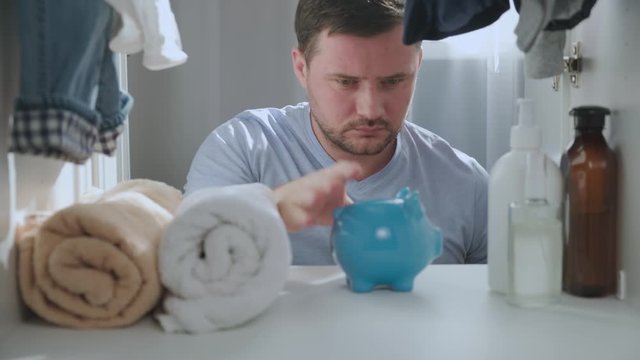 Caucasian unshaven man opens wardrobe in home and puts accumulated money coins and green currency in blue piggy Bank. Guy saves money for future, invests in investments for travel, mortgage or car.