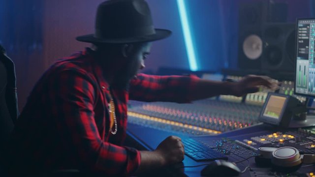Energetic Audio Engineer / Producer Working in Music Recording Studio, Mixing Tracks on Control Desk and Software to Create Hit Song Track. Black Artist Musician Enter His Studio at Sits at Workdesk