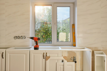 Cordless screwdriver and yellow building level on white kitchen furniture. Light interior, preparing to custom installation process, front view