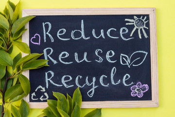 Chalk board Reduse Reuse Recycle sign on a yellow background with green leaves. Top view.