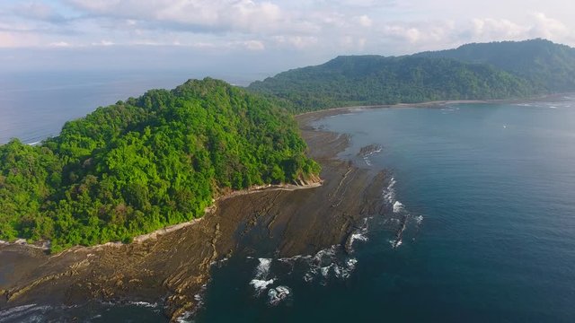 Side Strafe Flying Aerial View of Jicaron Island (Coiba Park) Water Edge with Slate Rock Cliff Shore and Dense Jungle Trees with Distant Islands