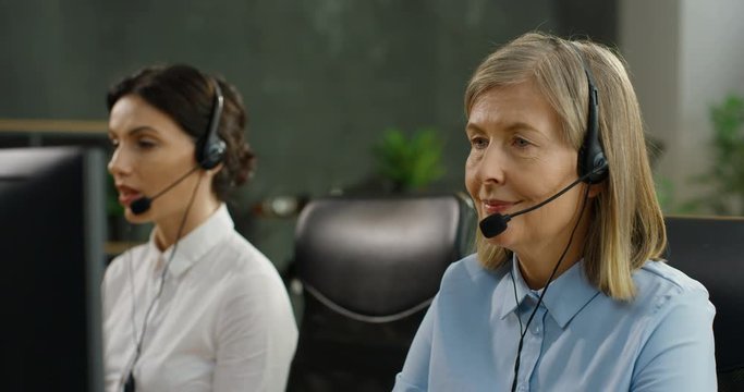 Close up of Caucasian senior woman in headset chatting with customer at computer and solving problem. Female colleagues in call center office. Young co-worker on background.