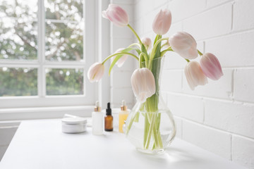 Close up of pale pink tulips in glass vase with skincare accessories in background on white table next to window (selective focus)