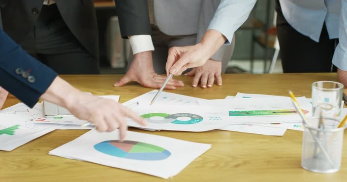 Close up of papers and documents with charts, graphics and diagrams lying on wooden table in office. Business meeting and discussion concept. Mixed-races female and male hands. Multiethnic partners.