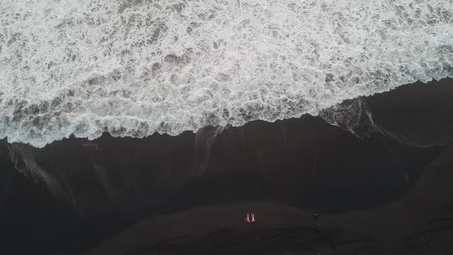 the camera flies vertically up from the beach with black sand and huge white ocean waves