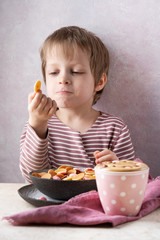 Trendy food - pancake cereal. Heap of mini cereal pancakes with chocolate drops in boul in kids hands. Little smiling baker boy tastes his portion. Funny food for kid during coronavirus