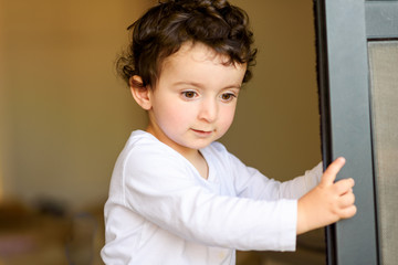 Funny boy with curly hair behind door at home. Photo of adorable young happy kid looking down. Selective focus.