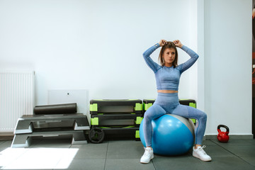 Fototapeta na wymiar Beautiful young caucasian girl in blue sports equipment ties her hair while sitting on a pilates ball in the gym