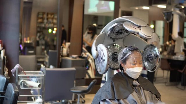 Korean woman wearing face mask and eye protection sits under heating lamps inside hair salon, static , hair perming, hair drying machine