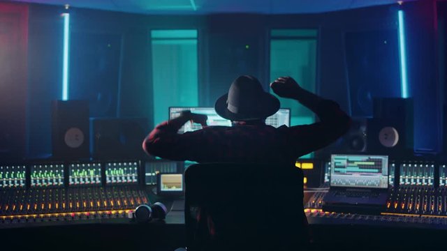 Artist, Musician, Audio Engineer, Producer in Music Record Studio, Uses Control Desk with Computer Screen showing Software UI with Song Playing. Celebrates Success with Raised Hands, Dances. Back View