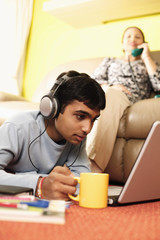 Young man using laptop on the floor, woman talking on the phone in the background