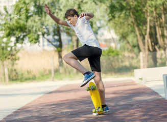 Teenager in white t shirt spends free time training skating in the city park. Practicing skills freestyle, making tricks. Cute nice boy enjoys sunny day outdoors with skateboard. Hobby, sport for kids