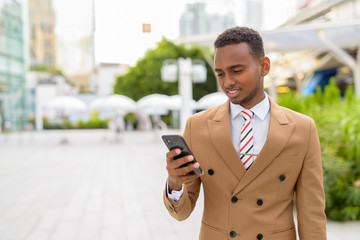 Happy young handsome African businessman using phone in the city outdoors