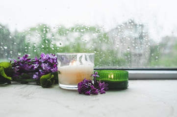 burning two candles white and green with branches of fresh purple blooming lilac sprig on the background of wet glass in drops and drips from the window