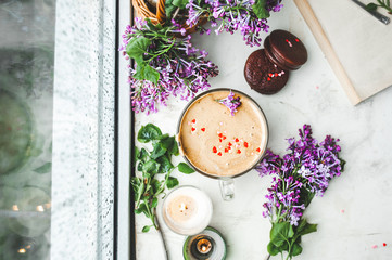 dalgona coffee with foam, chocolate cookies, burning candles and fresh lush purple-lilac flowers at the wet window with drops and drips from the rain. cozy beautiful Breakfast in bad  weather top view