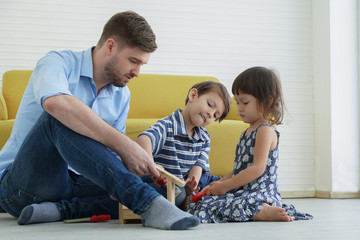 A Caucasian young father sitting on warm floor playing with happy little son and daughter construct with building wooden house blocks at living room at home