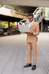 Fototapeta premium Full body shot of young African businessman using laptop in the city outdoors
