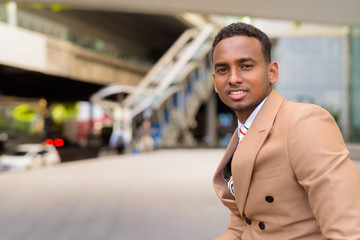 Happy young handsome African businessman smiling and sitting in the city outdoors