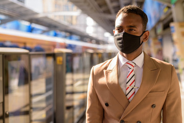 Young African businessman with mask thinking and waiting at the sky train station