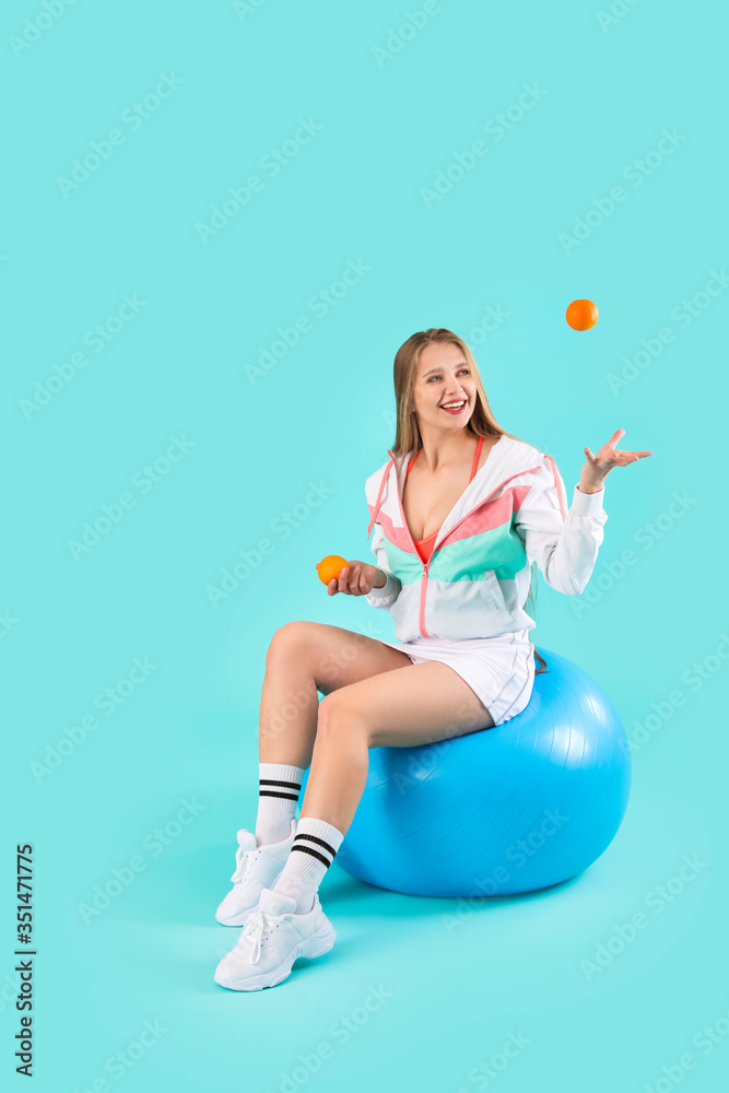 Wall mural Young woman with tangerines sitting on fitball against color background. Diet concept