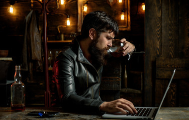 Fototapeta na wymiar Elegant man in leather jacket drinking a whiskey in bar. Brutal hipster man drinking alcohol. Stylish handsome bearded man enjoying whiskey and doing his business at laptop.