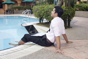 Young businessman relaxing by the pool with laptop on his lap