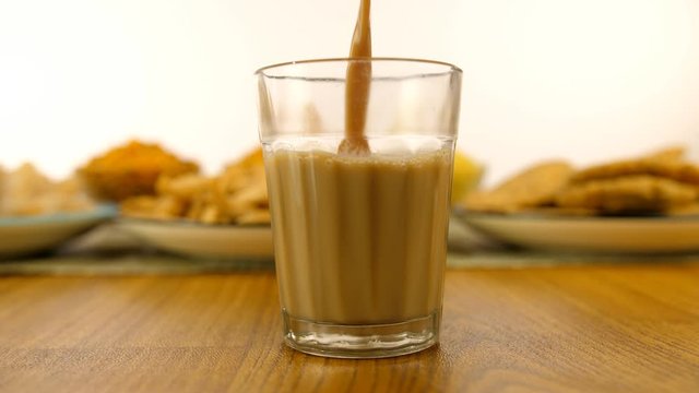 Hands pouring hot masala chai with plates of Indian snacks in the background. Closeup shot of hands serving tea in a transparent glass placed on a wooden table against the white background