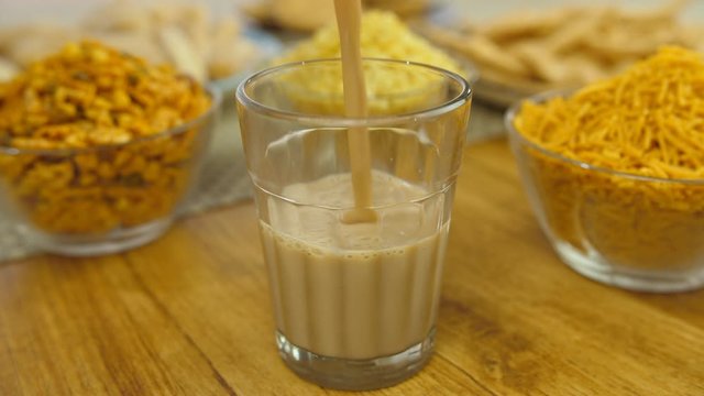 Closeup shot of hands serving hot masala chai in transparent glass - tea time. Morning tea table - Glass of tea surrounded by bowls of Namkeen mixture  Aloo Bhuja  and Moong Daal Namkeen