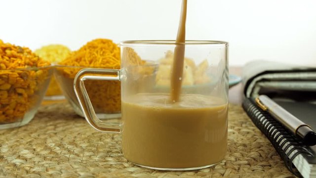 Closeup shot of hands serving hot masala chai in a glass cup with Indian snacks. Morning tea table with Namkeens  newspaper  a diary  a pen placed on a table mat - work from home during the lockdown
