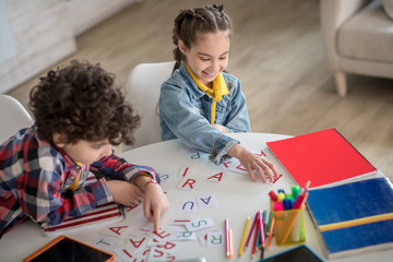 Curly boy and dark-haired girl sitting at round table, playing with alphabet letters, smiling