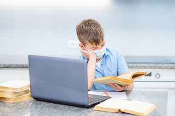 A boy in a medical mask is engaged at a laptop with a book. Distance education during illness