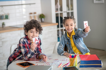 Curly boy and dark-haired girl sitting at round table, playing with alphabet letters, showing...
