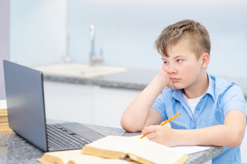 A boy in a blue shirt is reading a laptop with a book at home in the kitchen. The student laid his head in his palm and looks tired at the laptop camera. Quarantine Education