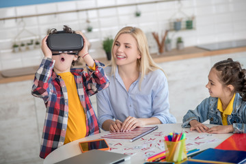 Curly boy and dark-haired girl sitting at round table with blonde female, boy wearing vr glasses, feeling excited