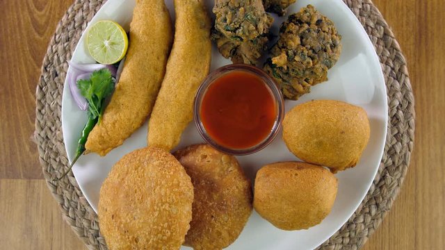 Zoom out shot of beautifully placed Indian snacks served with tomato ketchup . Top view of various starters like Mirchi pakora  palak pakora  paneer pakora  kachori  and red dip rotating on a turnt...