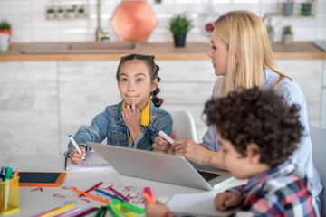 Curly boy and dark-haired girl sitting at table, blonde female helping them, explaining something to girl