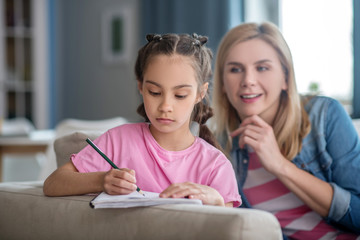 Dark-haired girl sitting on sofa, writing in her notebook, blonde female helping her, explaining something