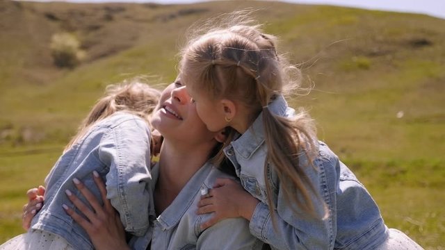 A happy young mother hugs her twin daughters on a field of green grass in Sunny weather, the daughters kiss their beloved mother. Slow motion.