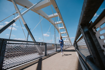 Naklejka premium A young beautiful caucasian girl in blue sports equipment is running on the bridge. Training in nature