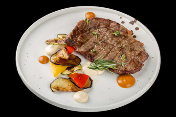 Grilled beef steak on a white plate. Served with a side dish of vegetables. Isolated on a black background.